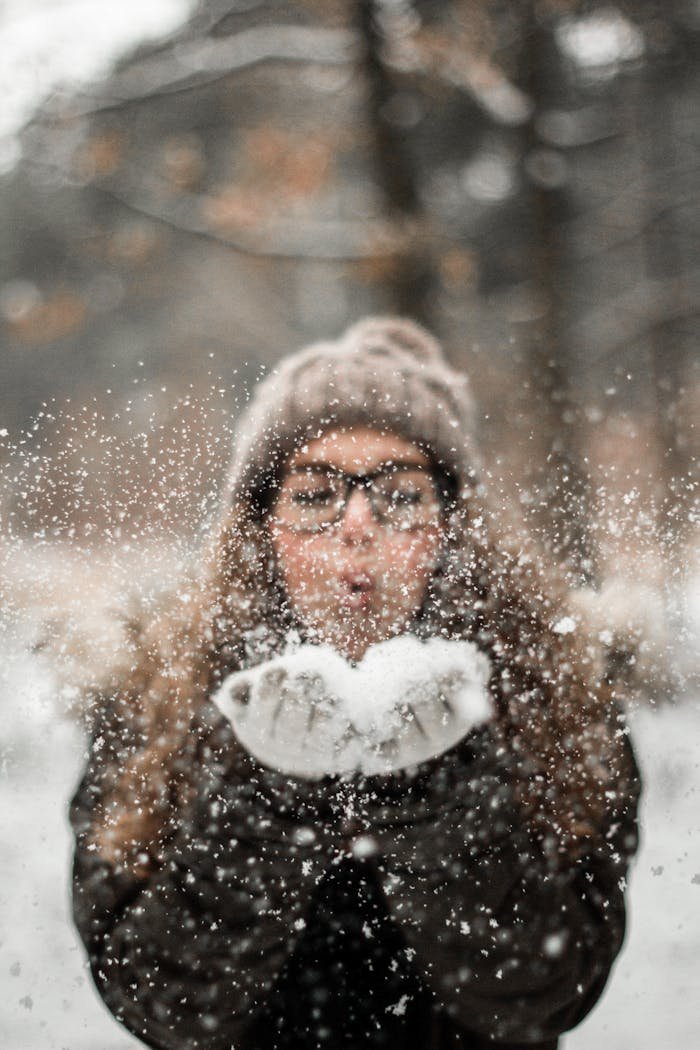 A woman in winter clothing playfully blowing snowflakes outdoors, capturing the joy of a snowy day.